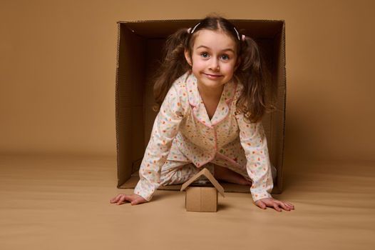 Beautiful child girl peeking out from behind a cardboard box, looking at camera. Craft carton box on a beige background with copy space for ads. Housing, insurance and real estate concept