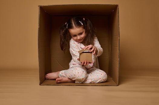 Adorable Caucasian child girl hiding inside a box and playing with a craft cardboard house model. Real estate, housing, insurance and social secure concept with copy space for advertisement