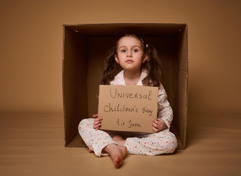 International Children's Day. Social advertising, information campaign for the fight for children's rights. Cute little girl with two ponytails holding cardboard poster sitting inside a cardboard box.