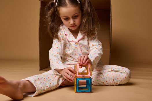 Cute adorable Caucasian baby girl sitting ahead a cardboard box and building a colorful house using square and triangle magnetic construction blocks. Fine motor skills development concept