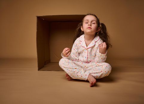 Charming kid adorable peaceful baby girl meditates against beige background, learning meditating practice.Life balance. Good vibes. Yoga training.Stay positive and optimistic. Copy space