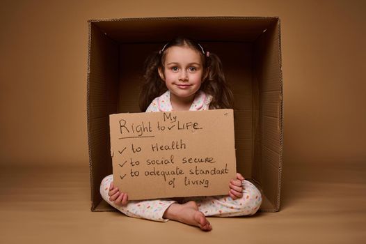 Happy smiling Caucasian little girl holding cardboard poster with social message, calling for respect for her rights, isolated over beige background with copy space. World Children's Day on July 1st.