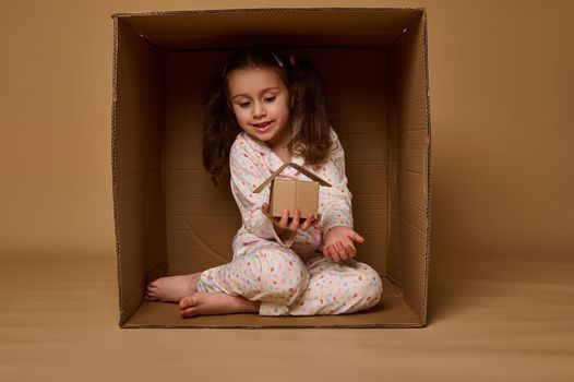 Adorable Caucasian little girl hiding inside a box and holding a craft cardboard house model. Real estate, housing, insurance and social secure concept with copy space for advertisement