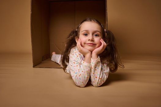 Smiling beautiful positive pleasant baby girlin pajamas with two ponytails inside a cardboard box, looking at camera isolated over beige background with copy space for ads