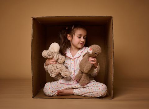 Charming little girl plays with her plush toys while sitting inside a cardboard box, isolated over beige background with copy ad space. Happy childhood, home, real estate and insurance concept