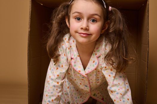 Close-up baby girl with two ponytails in pajamas peeking out from behind a cardboard box, isolated over beige background with copy space. Home, housing, insurance and real estate concept