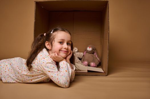 Beautiful Caucasian little girl, adorable child lying down on a beige background with copy ad space next to a cardboard box with a plush rabbit toy and a hard cover book