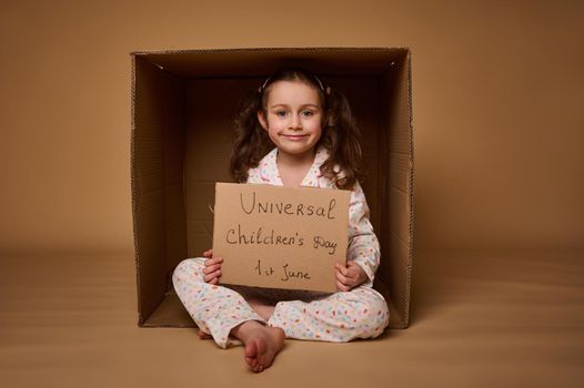 Beautiful little girl holding a cardboard banner, celebrating the International children's day on 1st July, sitting inside a carton box and smiles looking at camera, isolated on beige background