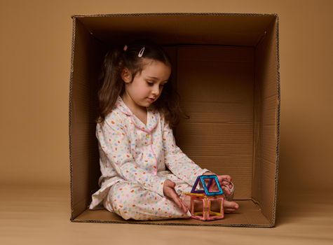 Adorable little girl in pajamas sitting inside a cardboard box and plying with a small colorful house buils from magnetic construction blocks.