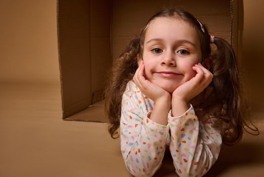 Headshot of a beautiful positive pleasant baby girlin pajamas with two ponytails inside a cardboard box, looking at camera isolated over beige background with copy space for ads