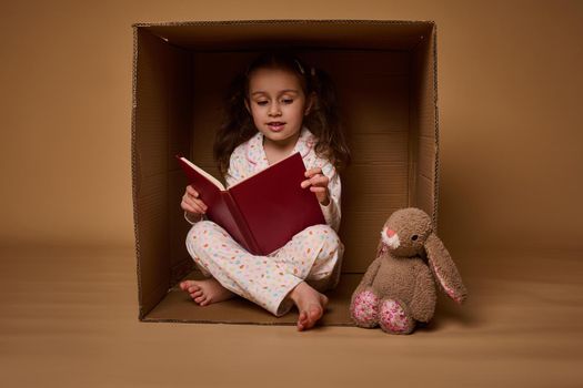 Smiling beautiful child, Caucasian positive pleasant baby girl in pajamas with two ponytails inside a cardboard box, looking at camera isolated over beige background with copy space for ads