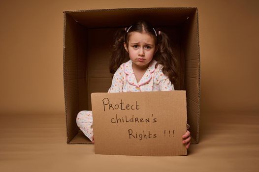 Upset little Caucasian girl holding a cardboard poster with social message, calling for respect for children's rights, isolated over beige background with copy space. World Children's Day on July 1st.