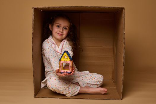 Little European girl holding a house built from magnetic square and triangle construction blocks, sitting inside a cardboard box and cutely smiling looking at camera, isolated over beige background