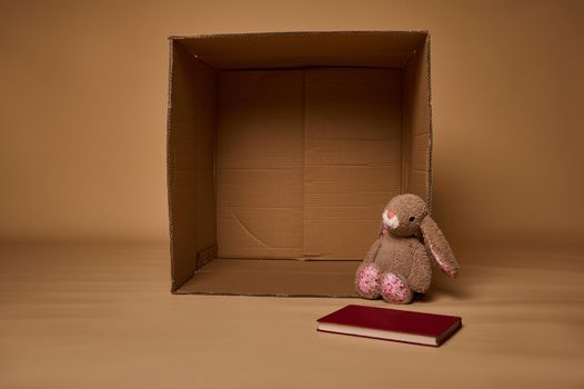 A plush rabbit toy and book next to an empty cardboard box, isolated over beige background with copy ad space