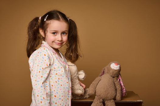 Close-up studio emotional portrait of a cute baby girl with two ponytails holding her plush toys and cutely smiles confidently looking at camera, isolated over beige background with copy ad space