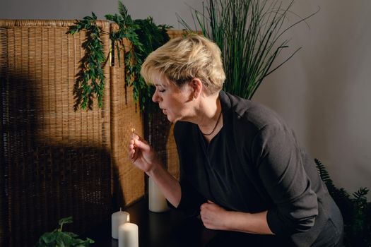 Portrait of a blonde, mature, concentrated and professional beautician, lighting scented candles with a hot wax massage matc.h. Relaxing atmosphere and soft lighting, decoration of plants and candles, massage table in the background and towels. Vertical