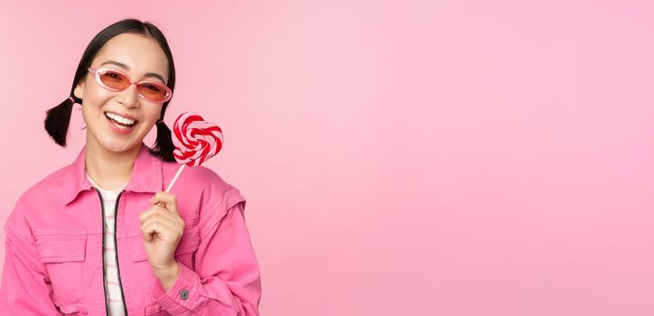 Stylish korean girl licking lolipop, eating candy and smiling, standing in sunglasses against pink background.