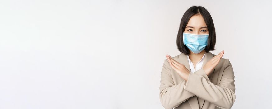 Coronavirus and workplace concept. Image of asian businesswoman, female in medical face mask shows stop, cross prohibit gesture, stands over white background.