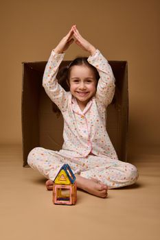 Beautiful child girl holds her hands above her head in the shape of a house roof, sitting inside a cardboard box while building house from magnetic constructor, smiles at camera over beige background