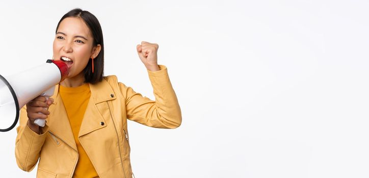 Portrait of young asian woman protester, screaming in megaphone and protesting, standing confident against white background.