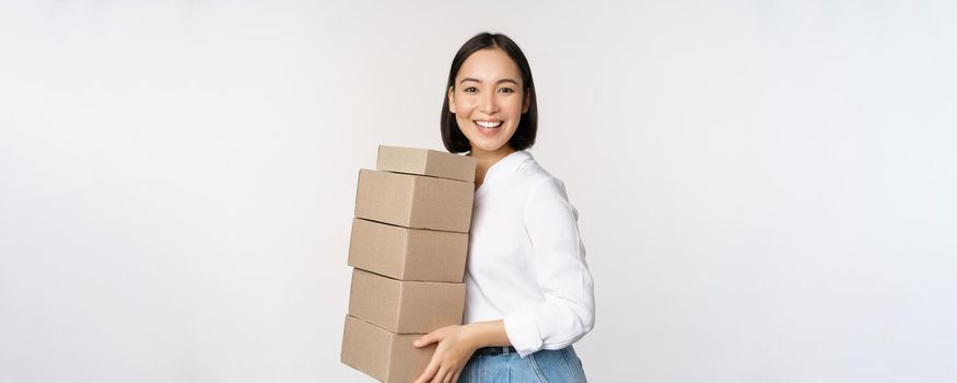 Image of smiling, happy asian woman holding pile of boxes, concept of delivery, shopping or business, white background.
