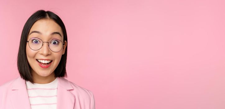 Close up portrait of asian businesswoman in glasses looking surprised at camera, amazed reaction, standing in suit over pink background.