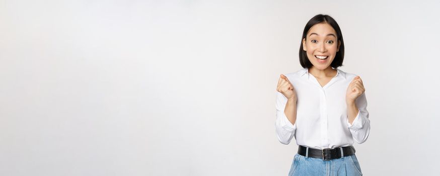 Enthusiastic asian woman rejoicing, say yes, looking happy and celebrating victory, champion dance, fist pump gesture, standing over white background.