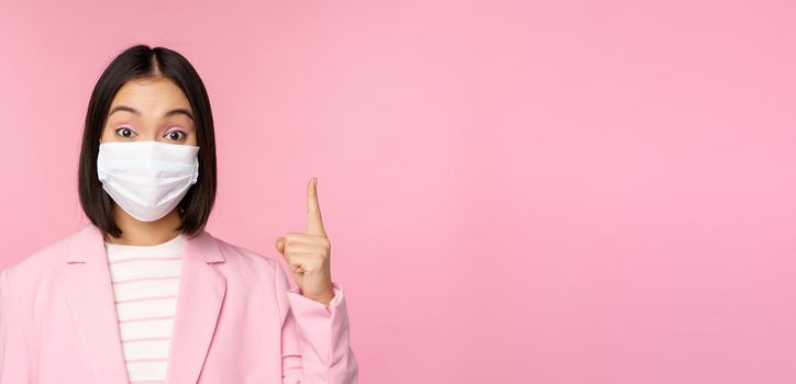 Close up portrait of asian businesswoman in medical face mask and suit, pointing finger up, showing advertisement, top banner, standing over pink background.