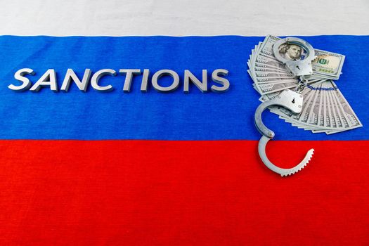 the word sanctions laid with silver metal letters on russian tricolor flag near dollar banknotes and handcuffs over it in linear perspective view