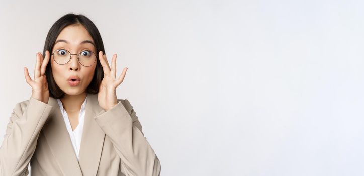 Portrait of asian businesswoman in glasses, looking surprised amazed at camera, standing in suit over white background.