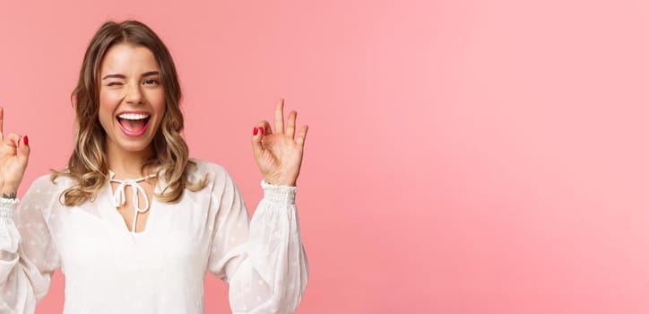 Close-up portrait of beautiful blond girl in white dress guarantee you will enjoy this event, show okay sign, confirm everything excellent, wink and smiling carefree, standing pink background.