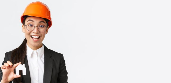 Close-up of professional broker in safety helmet and business suit selling homes. Cheerful architect showing minuature of house and smiling, working over new design or construction, white background.