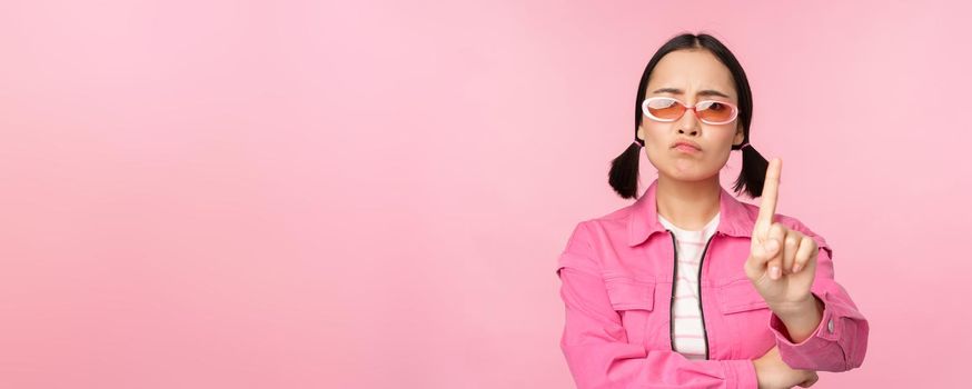 Image of serious, stylish asian girl in sunglasses, showing stop, prohibit gesture, taboo sign, saying no, standing over pink background.