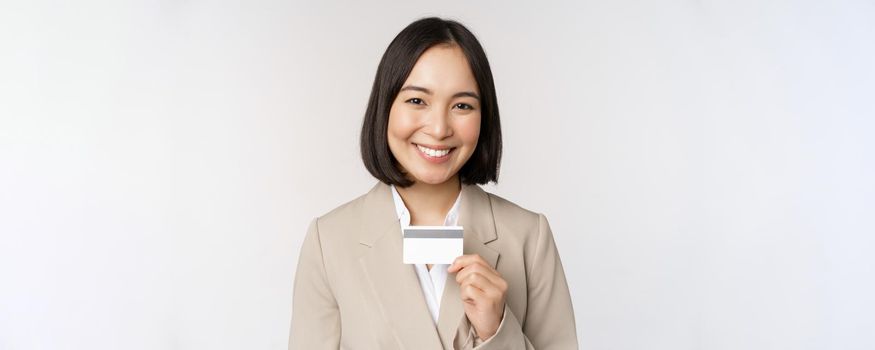 Smiling office clerk, asian corporate woman showing credit card, standing over white background in beige suit. Copy space