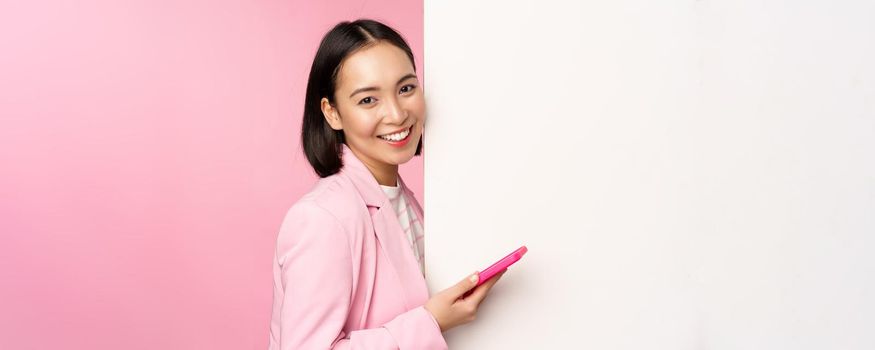 Image of korean female entrepreneur in suit, standing near info wall, advertisement on board, holding smartphone and smiling, posing over pink background.