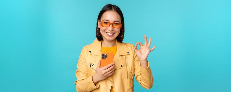 Smiling asian girl with smartphone, shwoing okay, ok sign in approval, standing over blue background. Copy space