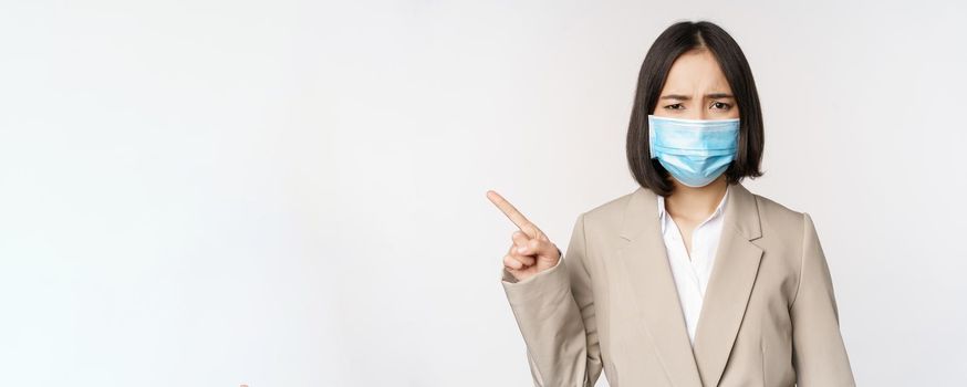 Coronavirus and work concept. Frustrated asian business woman in medical face mask, pointing finger left and complaining, standing over white background.