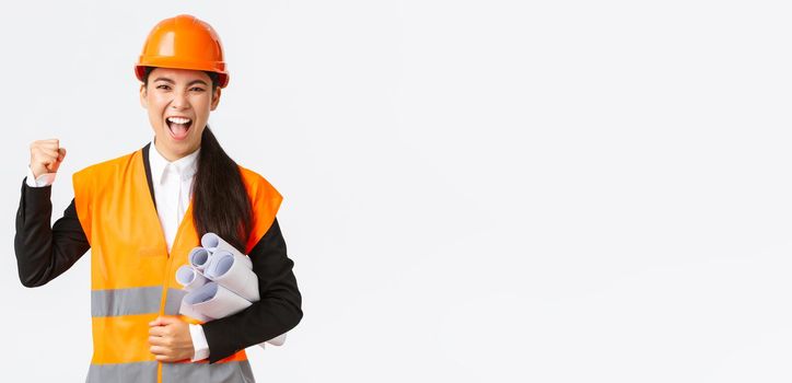 Successful female asian engineer, architect in safety helmet and reflective jacket carry blueprints of building project and fist pump in celebration, shouting yes, winning tender, white background.