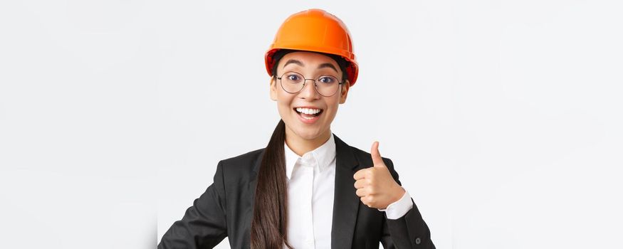 Close-up of professional pleased asian female chief engineer, architect in business suit and safety helmet showing thumbs-up in approval, give permission, recommend construction company.