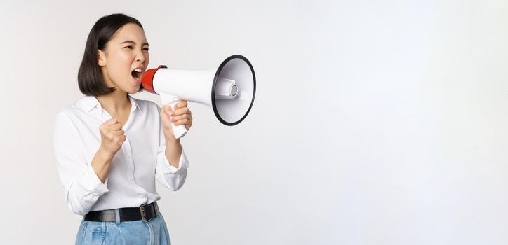 Enthusiastic asian woman, girl activist shouting at protest, using megaphone, looking confident, talking in loudspeaker, protesting, standing over white background.