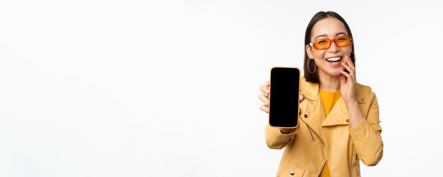 Enthusiastic asian female model, showing smartphone app interface, online store or website on mobile phone screen, standing over white background.