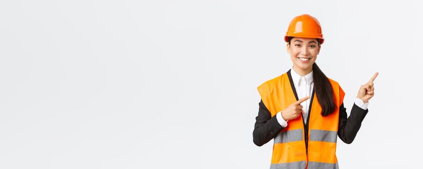 Building, construction and industrial concept. Smiling female asian engineer in safety helmet and reflective clothing showing object, pointing fingers right, saleswoman introduce estate to client.