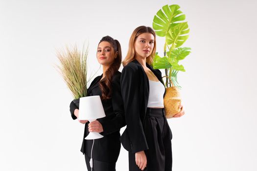 Two business woman dressed black suit holding houseplant and lamp standing isolated on white background. Moving concept. Relocation new office with positive. Caucasian female business person moving