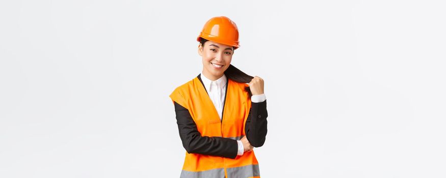 Portrait of confident smiling asian female construction manager, engineer at building area, wearing safety helmet and reflective jacket looking delighted at project, being pleased with finished work.