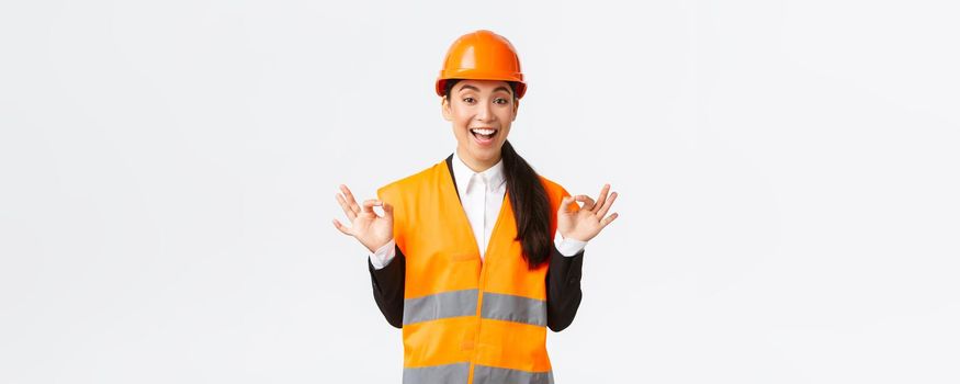 Excited and satisfied asian female chief engineer pleased with amazing work, showing okay gesture, assure clients, guarantee perfect quality, standing in safety helmet over white background.