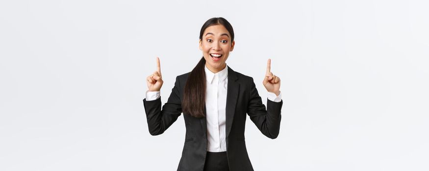 Excited smiling asian saleswoman in suit suggest great deal, pointing fingers up as telling details. Businesswoman making announcement and showing top banner, white background.