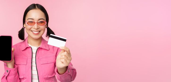 Image of smiling korean woman showing credit card and mobile phone screen, smartphone application interface, paying online, shopping contactless, standing over pink background.