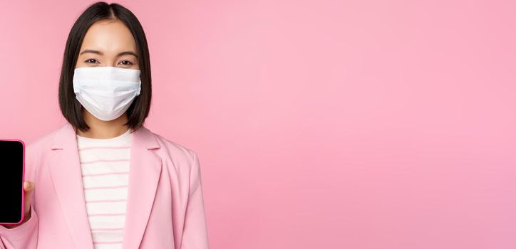 Portrait of smiling korean saleswoman in medical face mask, business suit, showing smartphone screen, standing over pink background.