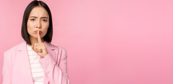 Hush, taboo concept. Portrait of asian businesswoman showing shush gesture, shhh sign, press finger to lips, standing over pink background in suit.