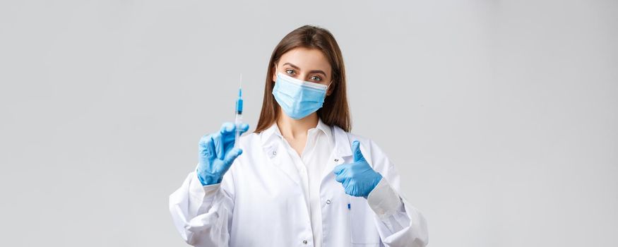 Covid-19, preventing virus, healthcare workers and quarantine concept. Determined young doctor in medical mask and rubber gloves, scrubs show thumbs-up and syringe with coronavirus vaccine.